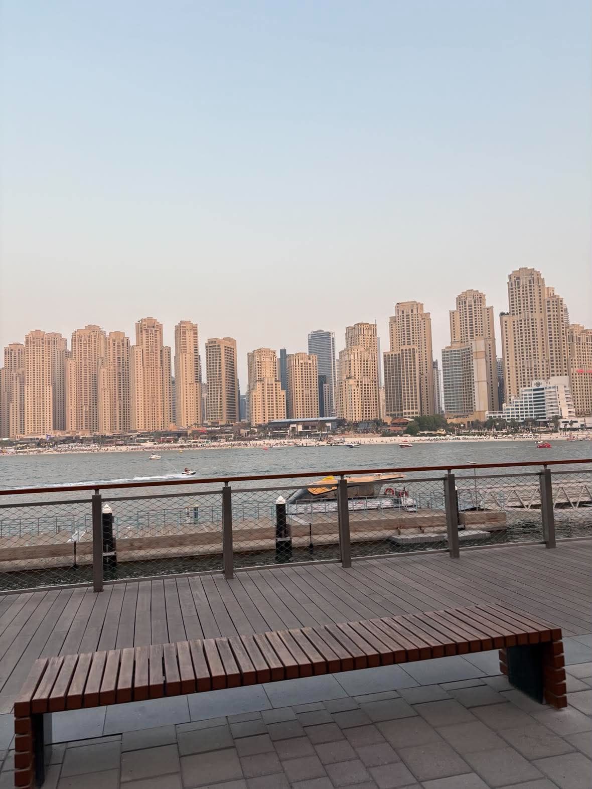 The JBR towers and the beach seen across the water from the Bluewaters boardwalk, a wooden bench in the foreground