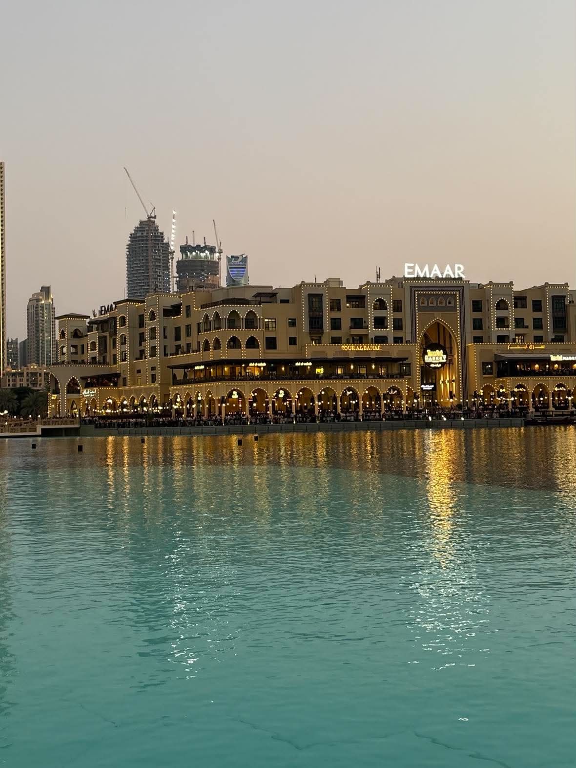 Souk Al Bahar's arched, lit facade and terraces reflected in Burj Lake at dusk, with cranes on unfinished towers behind